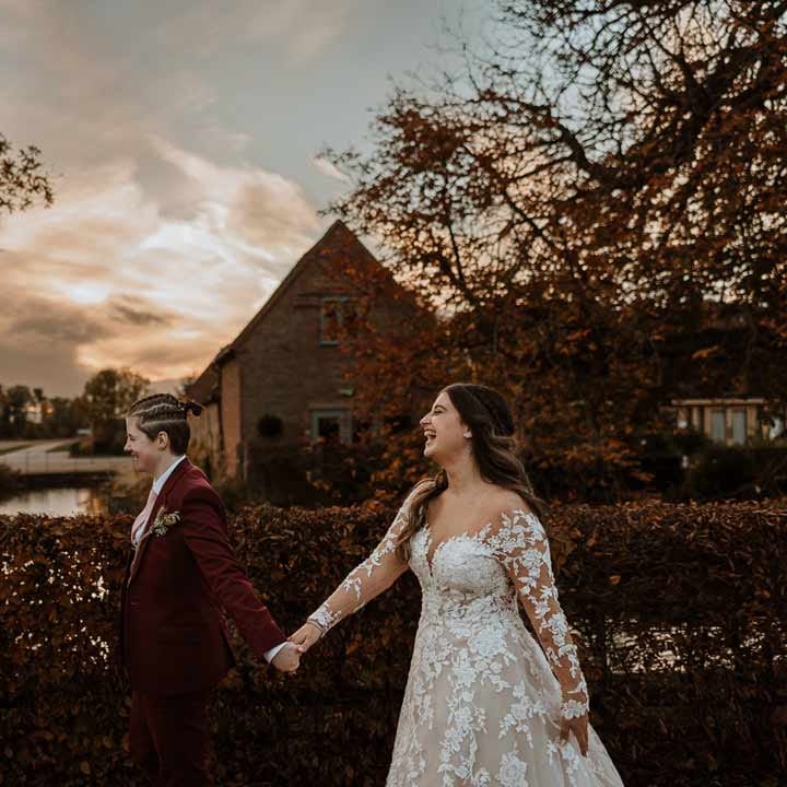 Bride in maroon bridal suit with grey waistcoat, dusky pink tie and autumnal dried flower boutonniere walking hand in hand with bride in long sleeve lace illusion sleeve wedding dress with overlay