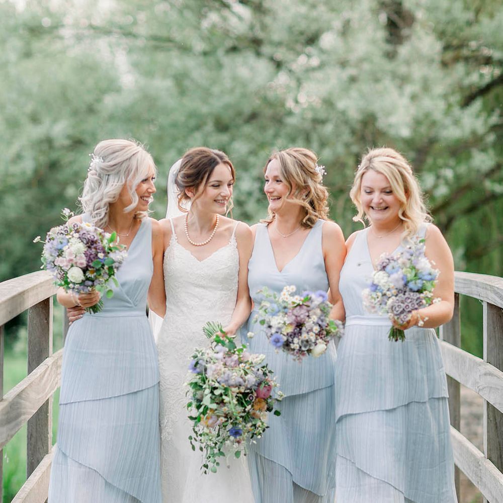 Bride in strappy white wedding dress walking over bridge with bridesmaids wearing sleeveless layered blue bridesmaid dresses 