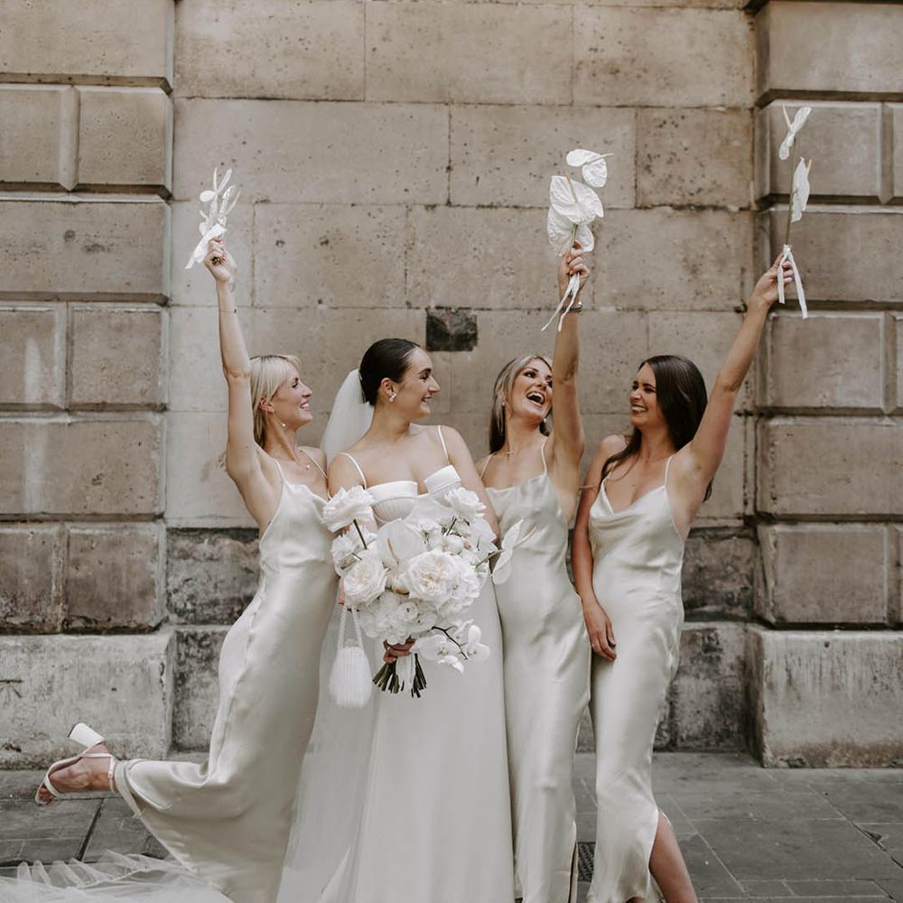 Bride in off the shoulder satin wedding dress standing with bridal party in sleeveless satin white bridesmaid dresses 
