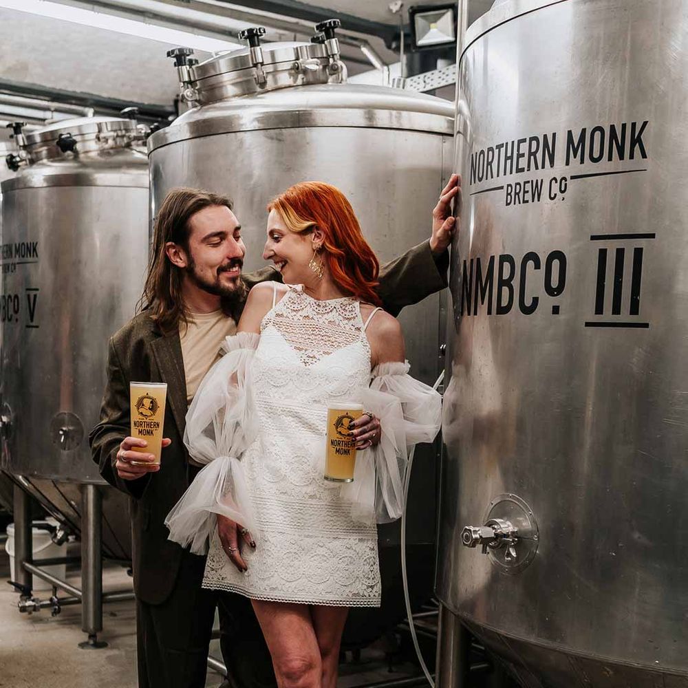 Bride wearing 70s inspired boatneck short lace wedding dress with detachable tulle puff sleeves and platform gold wedding heels standing by beer fermenter with groom wearing deep brown suit with off white t shirt and vans at Northern Monk Refectory Leeds