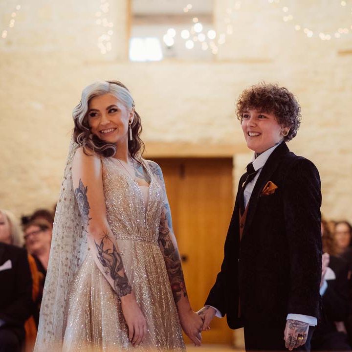 Bride in dark velvet bridal jacket with rust waistcoat and pocket square, black suit trousers and gold pocket watch holding hands with bride in sleeveless deep v neck sparkly gold wedding dress with sheer and gold polka dot wedding veil at the alter of Kingscote Barn Tetbury 