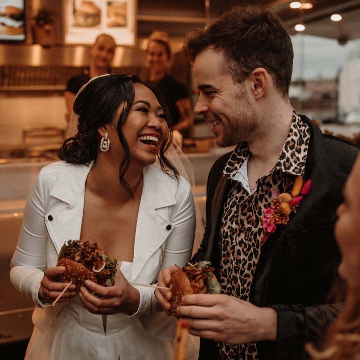 Bride in white leather jacket having wedding burger with groom in wet look black grooms suit with leopard print shirt and pink and orange boutonniere