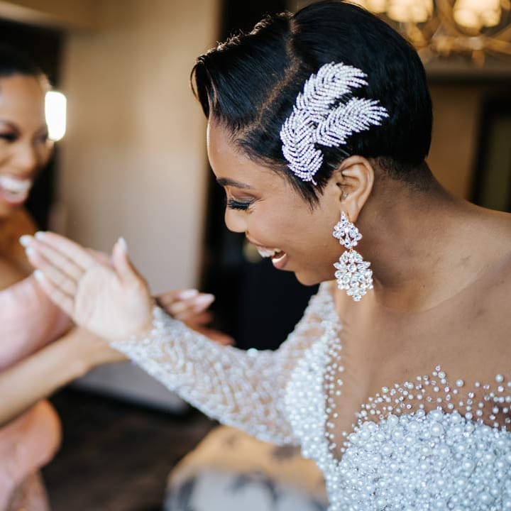 Bride in illusion v neck sheer off shoulder long sleeve pearl embellished wedding gown, large silver bridal hair accessory and silver bridal jewellery high fiving bridesmaid in pink off shoulder bridesmaid dress