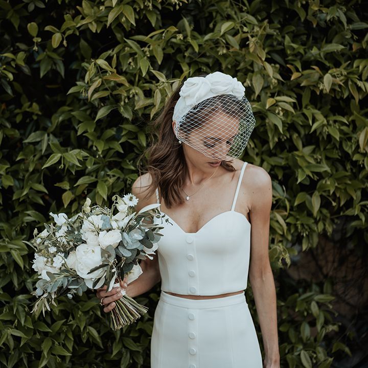 bride in separates with headband and blush veil holding a white flower and green foliage wedding bouquet 