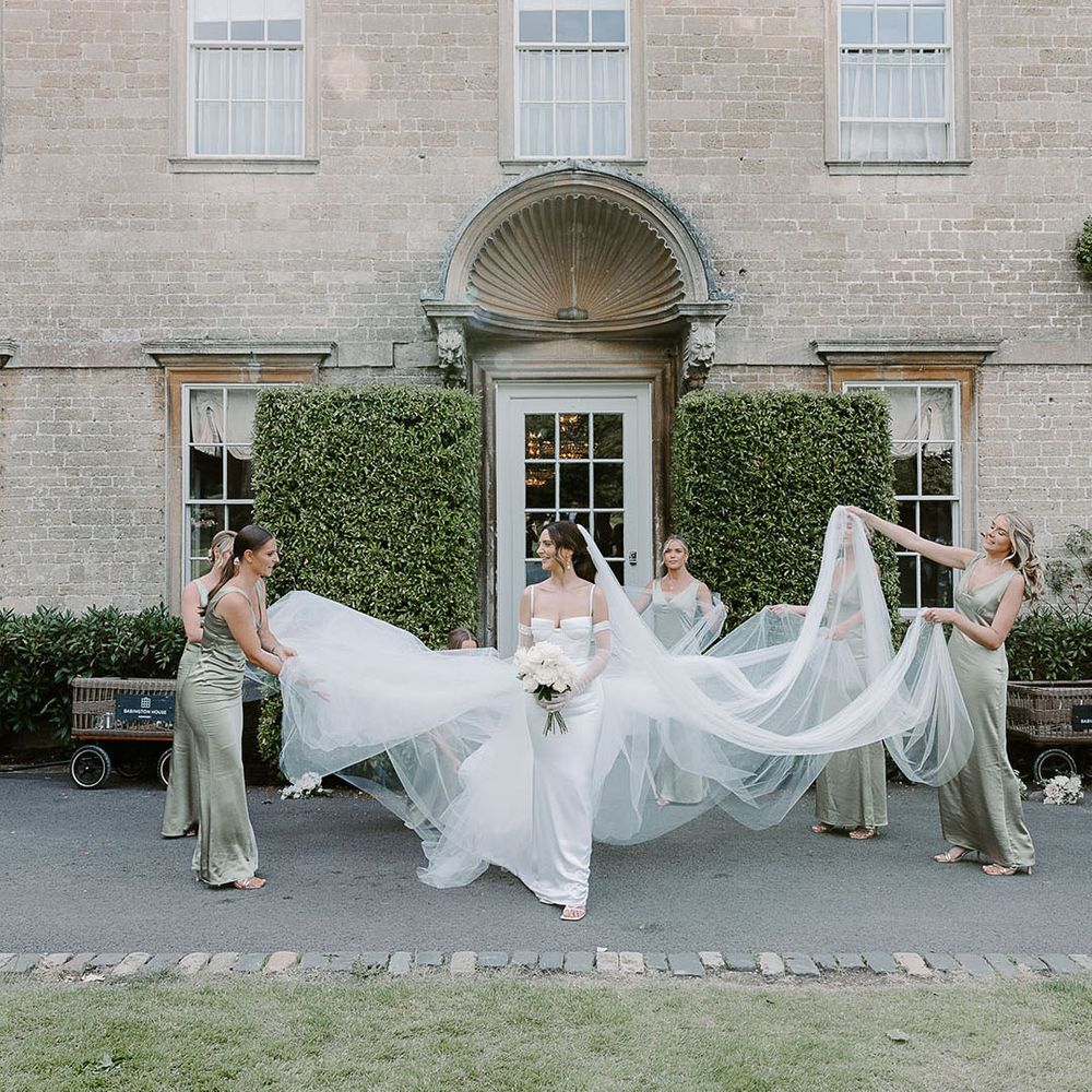 Bridal party in green satin bridesmaid dresses holding her veil and overskirt 