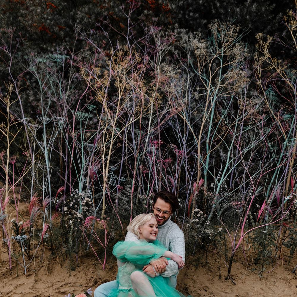 Groom in a grey jumper and stone wash jeans embracing his bride in a sheer wedding dress on the beach 