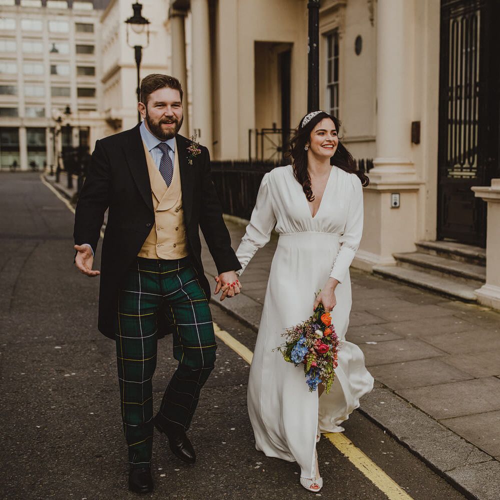 Bride and groom walking through London, groom wears green and yellow tartan trousers and three piece suit, bride wears long sleeved silk dress with front slit