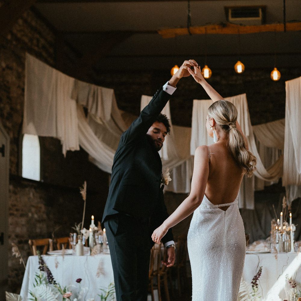 Stylish bride in a low back sparkly wedding dress with sleek ponytail being twirled by her husband in their reception room 