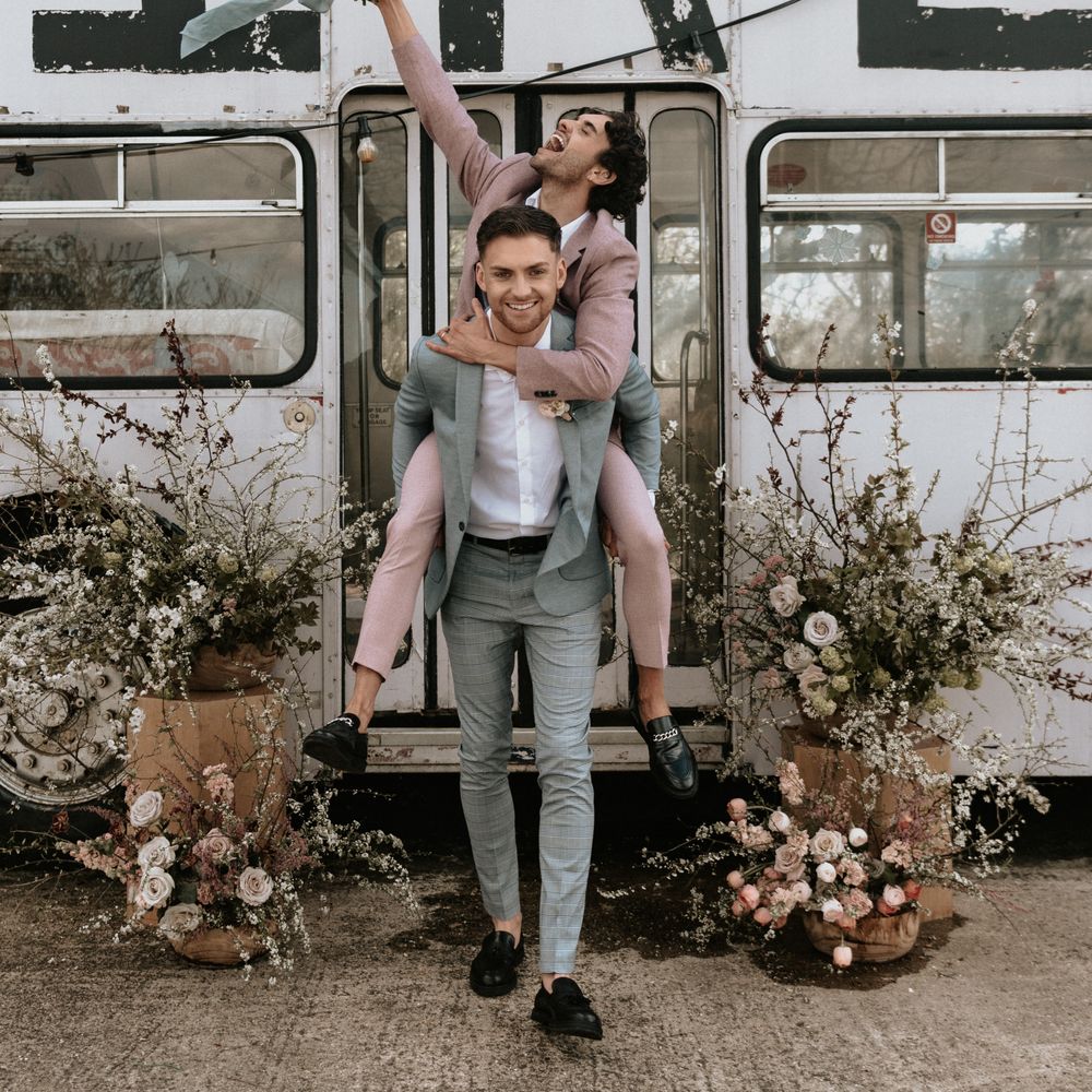 Groom carries groom on his back whilst both wearing pastel suits