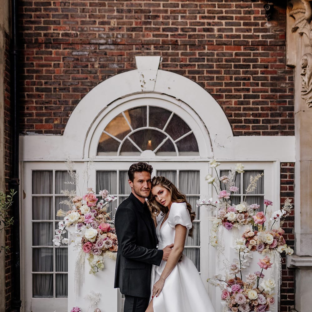 Stylish bride and groom in an all black suit and tie standing at the altar with pink floral arrangements 
