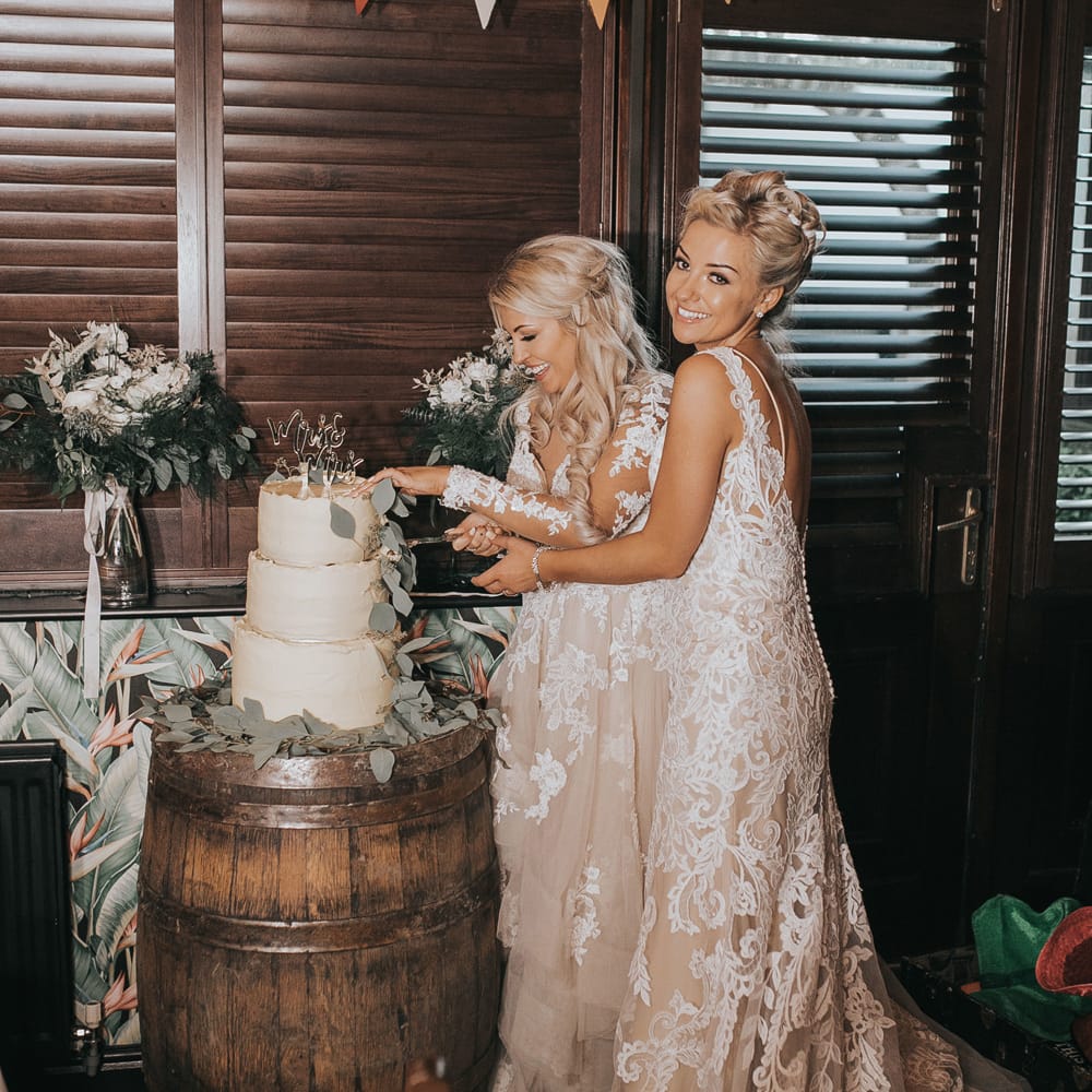 Two brides in white dresses cut a three tiered white wedding cake at Crab & Lobster Fairytale Wedding.