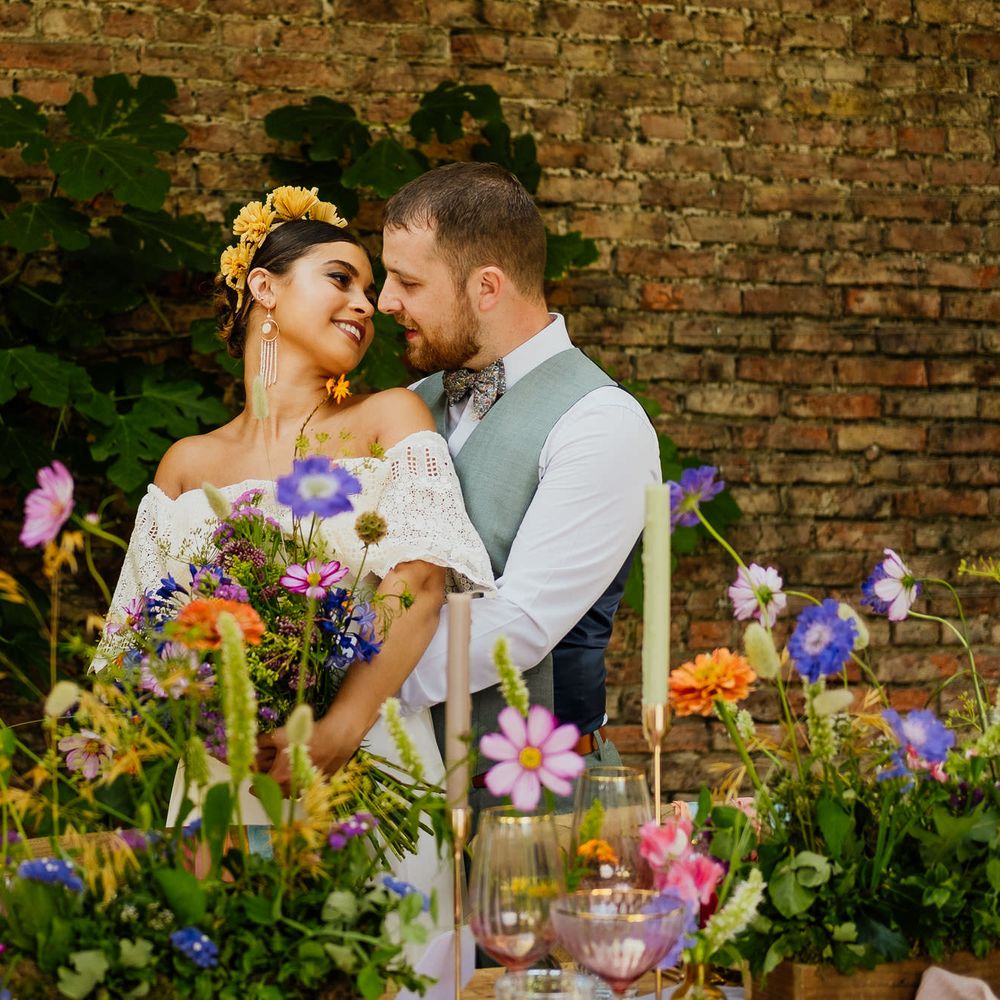 Groom embracing his boho bride in a strapless wedding dress and yellow headband standing behind a table full of colourful wildflowers and candles 