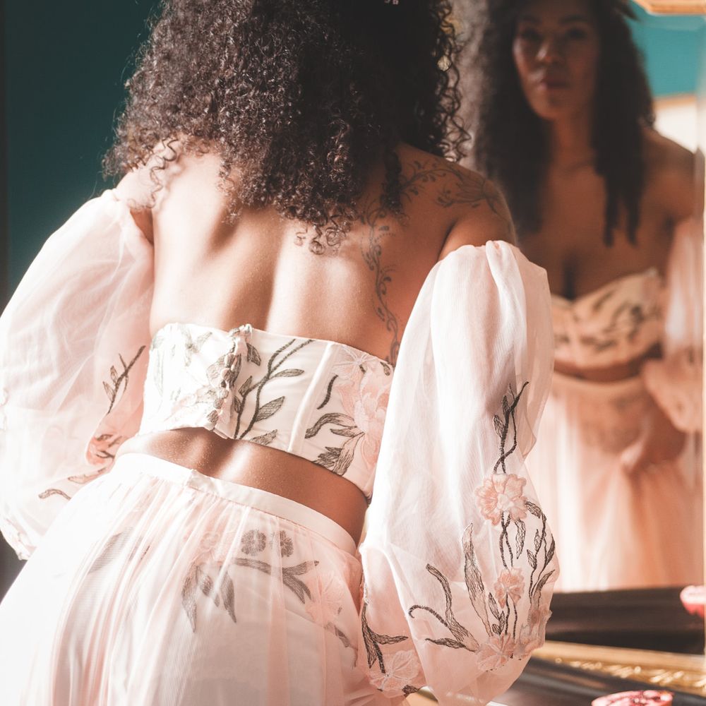 Black bride with long curly hair in blush pink bridal separates with appliqué detail looking in the mirror