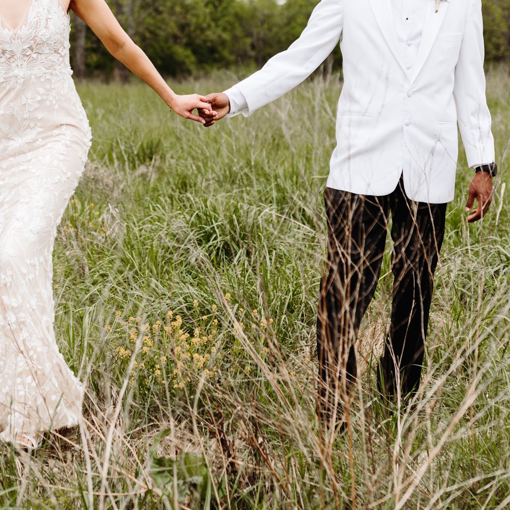 Bride in white embroidered Made With Love wedding dress holds hands with groom in white tuxedo jacket and black trousers in field after backyard wedding