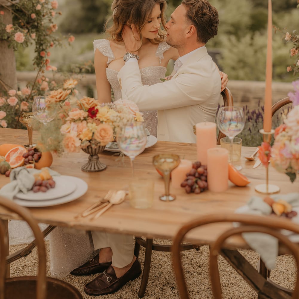 Groom in a stone coloured suit embracing his brides face at their outdoor wedding reception with orange candles, gold accents and blush flowers 