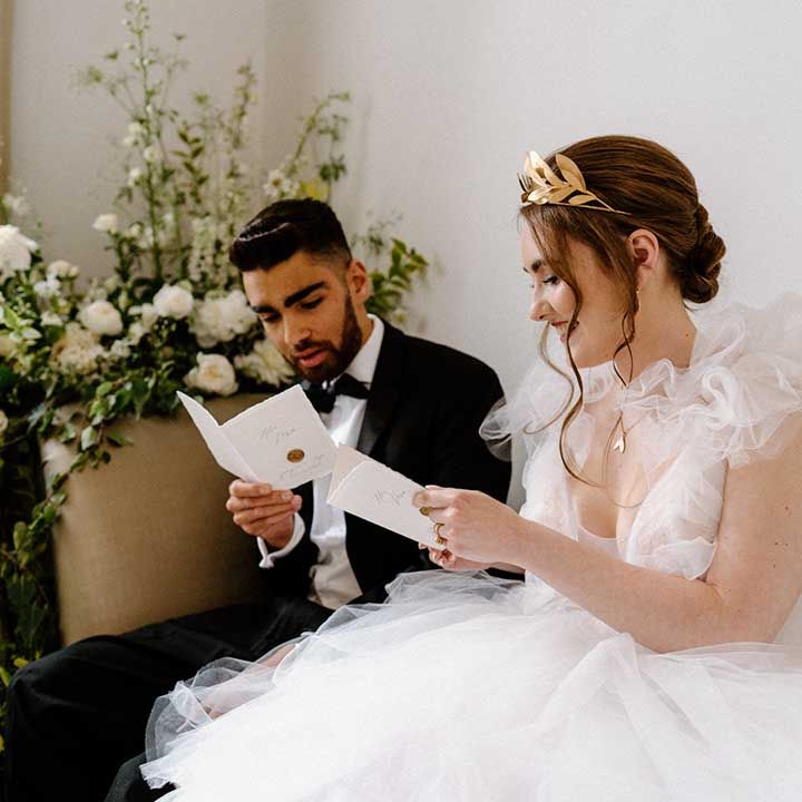 Bride in a fluffy tulle wedding dress with gold laurel crown sitting on a chaise lounge with her groom in a tuxedo 