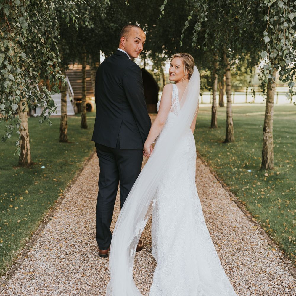Bride in white lace Justin Alexander wedding dress and veil holds hands with groom in dark suit as they both look over their shoulders in the grounds at Tythe Barn wedding with barn wedding flowers