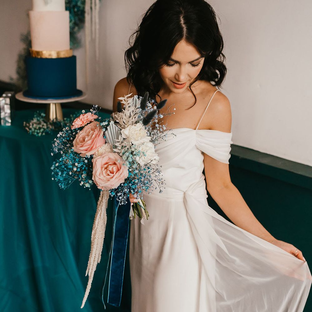 Bride with shoulder length wavy hair in a cold shoulder wedding dress holding her pink rose and teal gypsophila wedding bouquet tied with velvet ribbon 
