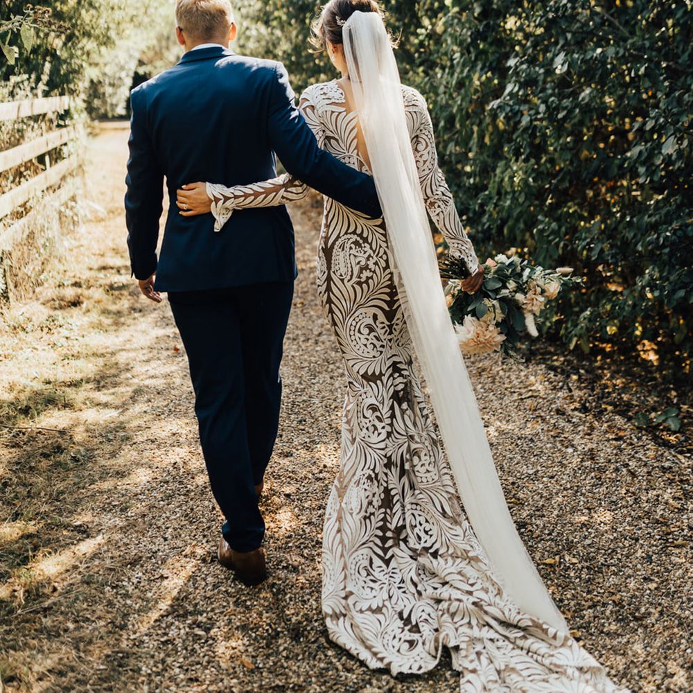 Bride in long Boho wedding dress with veil walking in forest setting with Groom in Navy suit 