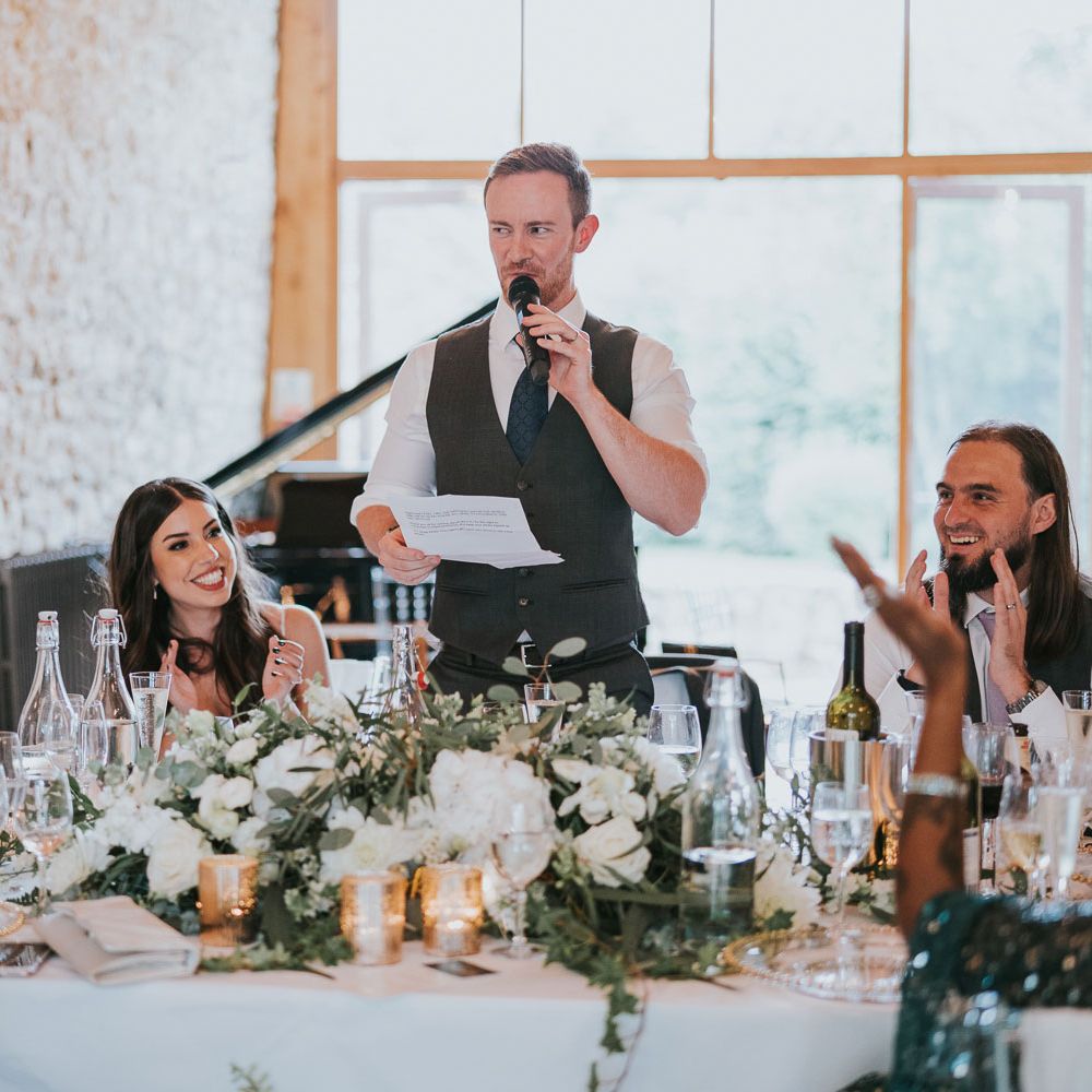 Groom in white shirt and grey waistcoat makes speech from round top table at wedding reception in Buckinghamshire