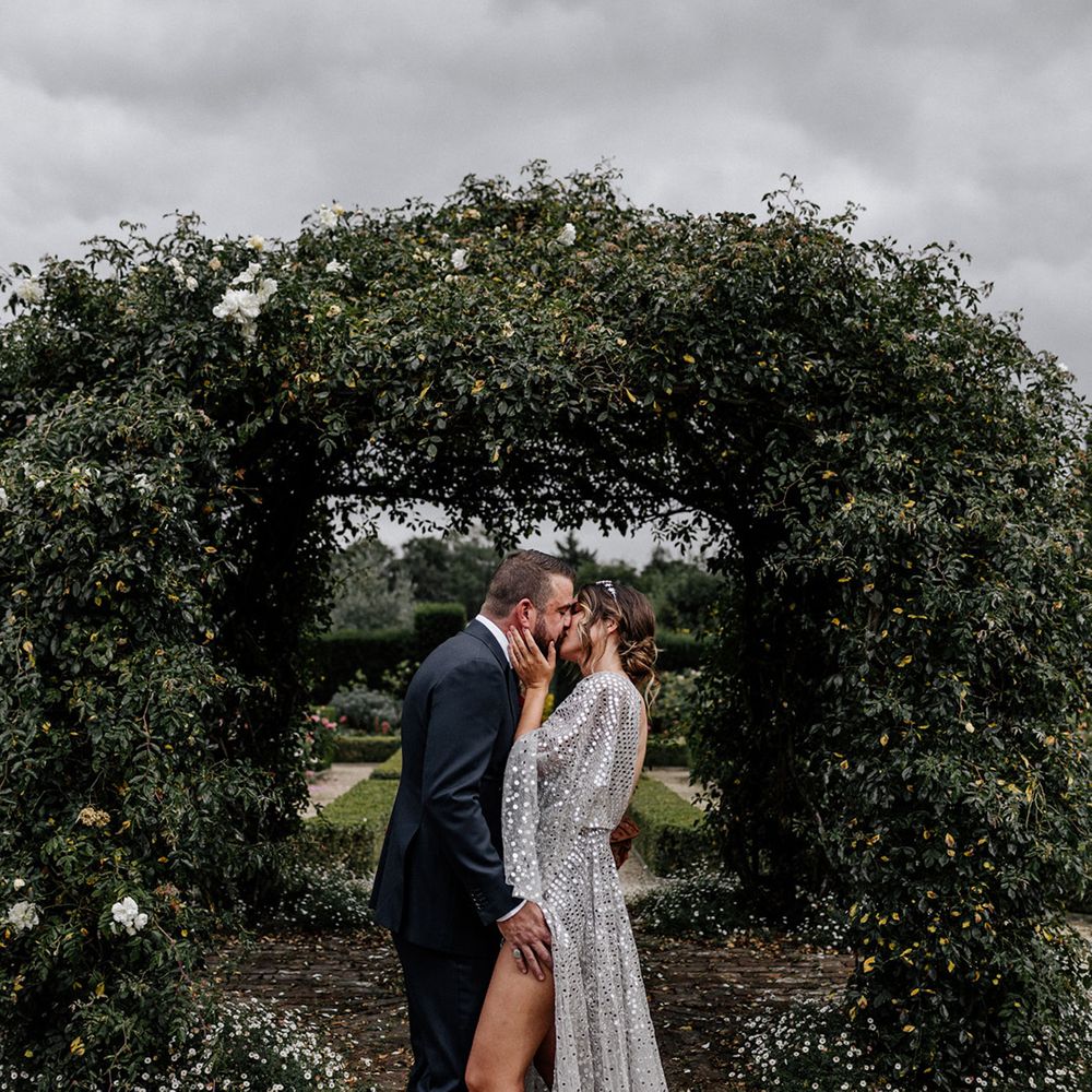 Bride in a silver sequin wedding dress and groom in a navy suit kissing in front of a greenery arch 