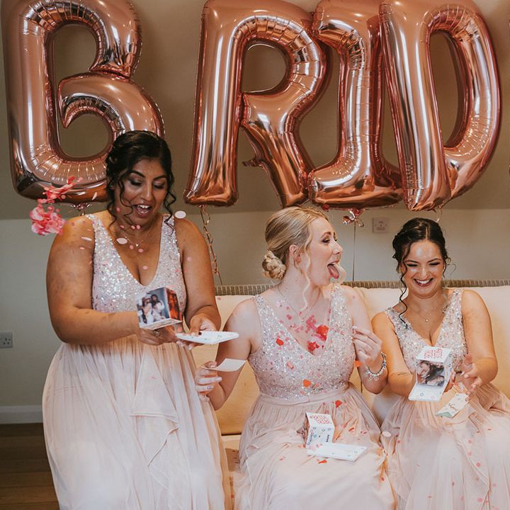 Bridesmaids in matching pale pink sparkly dresses opening gifts in front of rose gold balloons