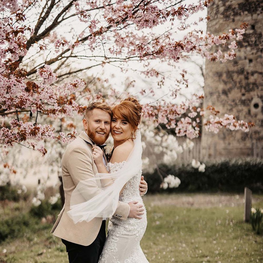 Bride and groom embrace under blossom trees for wedding in spring at rustic barn venue