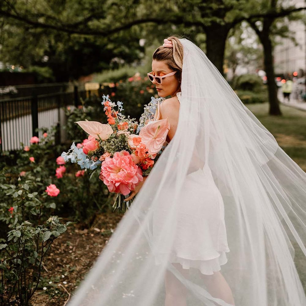 Bride in a long veil and sunglasses holding a coral pink and blue wedding bouquet with peony, anthuriums and delphiniums