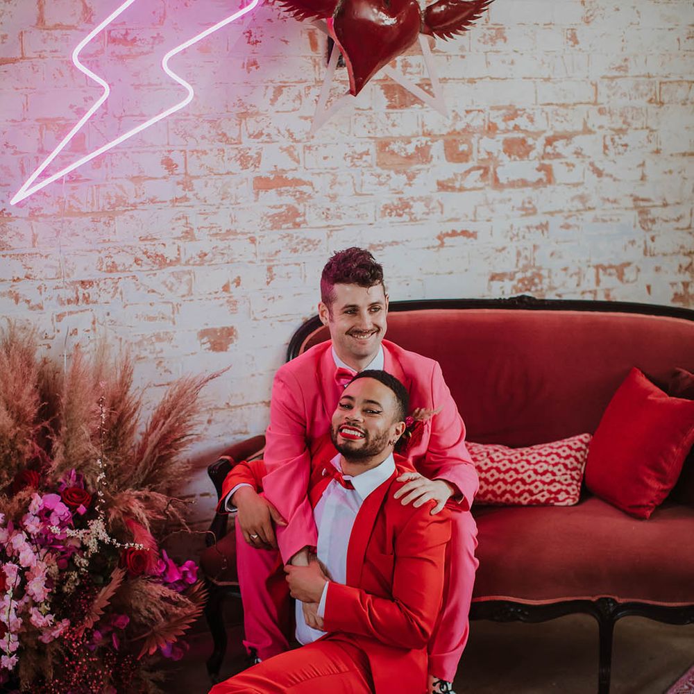 LGBTQI+ couple at Prestwold Hall Barn wedding sitting on a velvet couch surrounded by quirky neon sign and heart wedding decor 