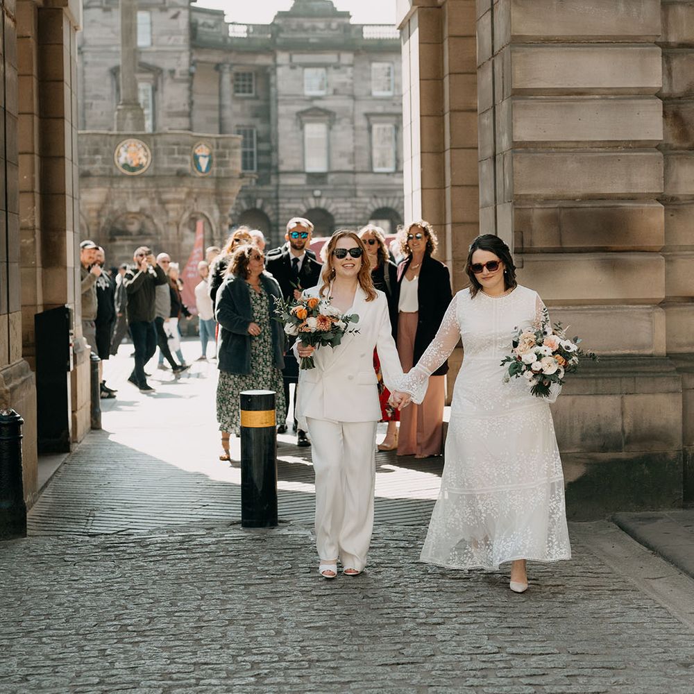 Two brides walk with one another whilst wearing sunglasses and carrying bouquets filled with white and orange florals 