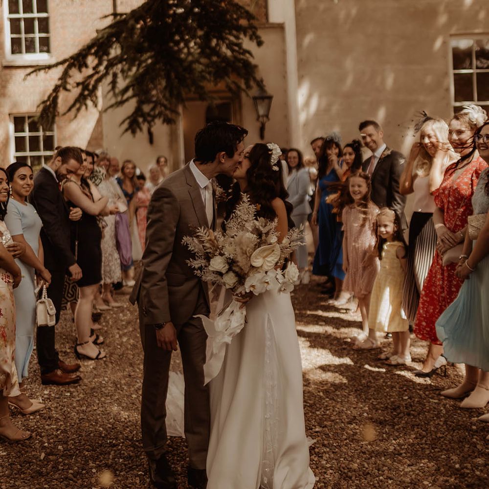 Bride and groom share a kiss together with bride carrying white wedding bouquet with dried grass as wedding guests watch 