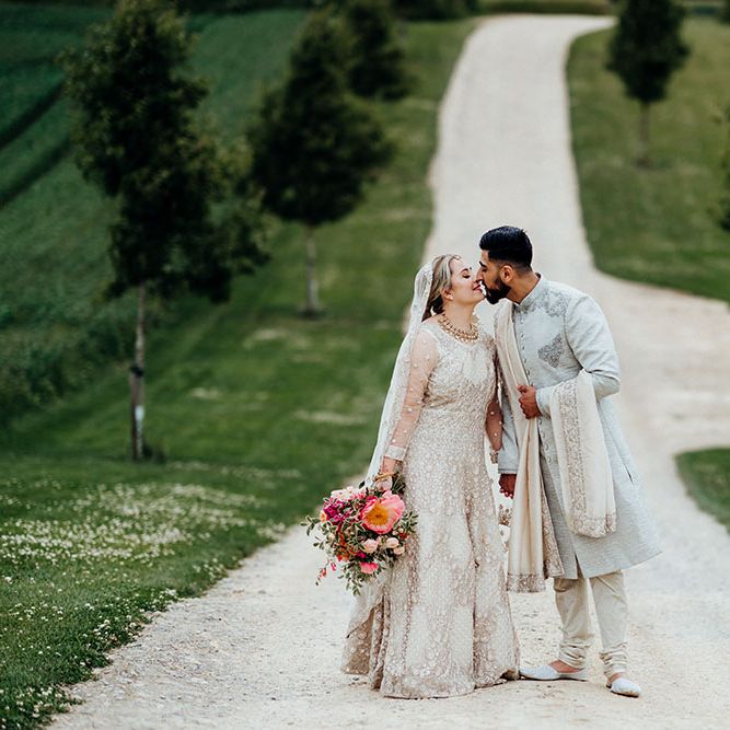 Bride in traditional white embellished saree holds bright floral bouquet and leans in to kiss her groom who wears embellished sherwani 