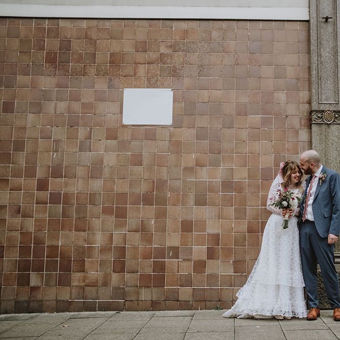 Bride carries sustainable pink and white floral bouquet whilst stood with her groom who wears grey suit with pale pink tie 