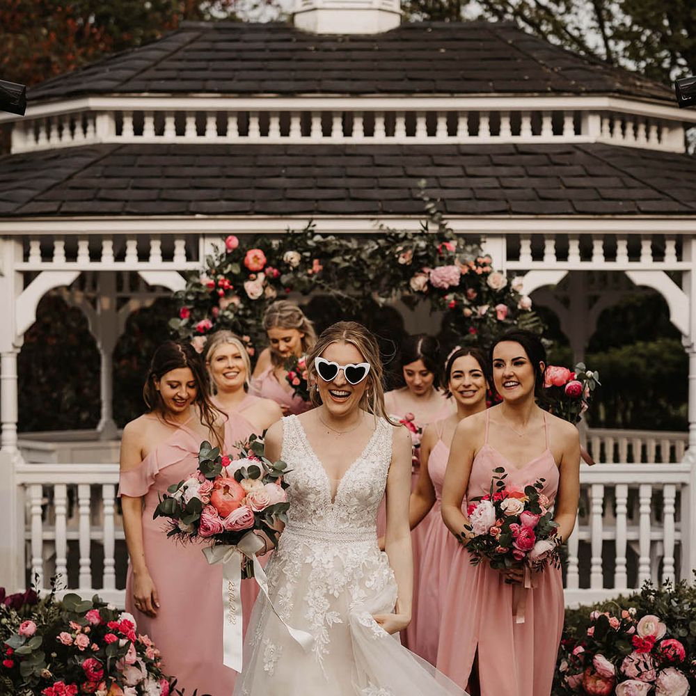 Bride in retro cat eye sunglasses in lace wedding dresses with bridesmaids in pink dresses in front of the pink flower arch