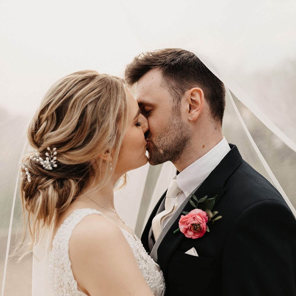 Groom in a dark morning suit and pink flower buttonhole shares a kiss with the bride 