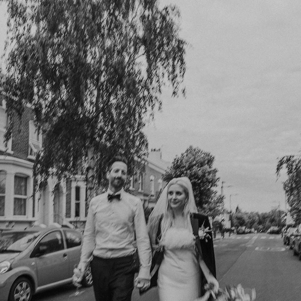 Bride in short satin dress with feather trimmed neckline and groom's blazer and groom in white shirt with bowtie holding hands and walking at Hackney Studios wedding