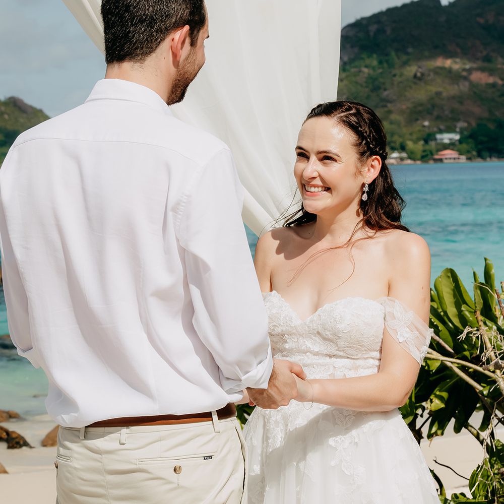 Bride beaming at groom and holding his hand wearing off the shoulder lace wedding dress with lace floral details and cathedral length veil holding white tropical bouquet 