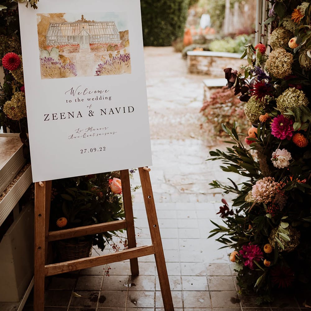 Wedding welcome sign with minimal type and watercolour painting stood on wooden easel by large floral wedding arch with white and pink carnations, garden roses, red strawflower, eucalyptus, baby’s breath and various dried flowers 