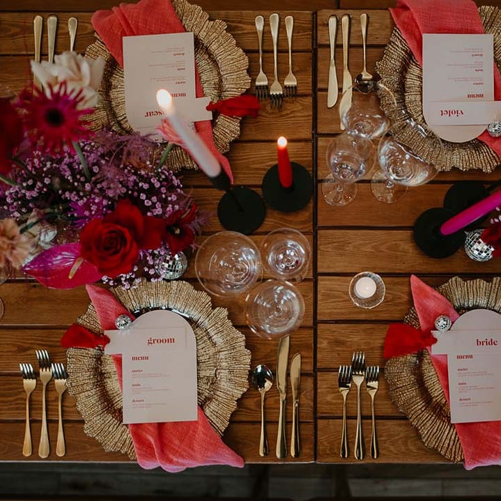 Four round gold charger plates with gold cutlery, red, white and pink tapered candles in black candlestick holders, bright pink napkins, retro wedding menus, disco ball wedding decorations and bright pink, magenta and purple floral arrangements 