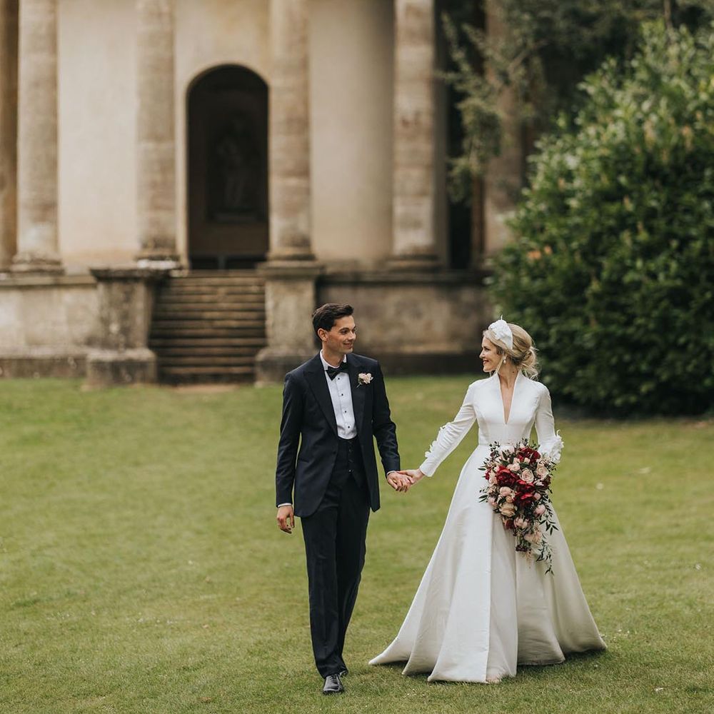 The groom in black tie and bride in a conservative long sleeve wedding dress walk hand in hand around the grounds at Stowe House 