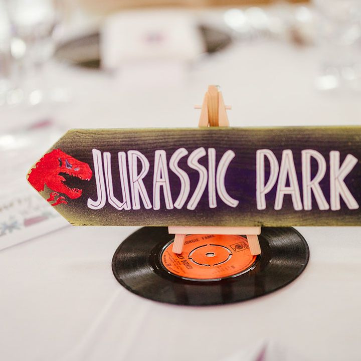 Quirky wedding table number sign in the style of Jurassic Park; cream arrow sign with "Jurassic Park" lettering on top of miniature vinyl record 