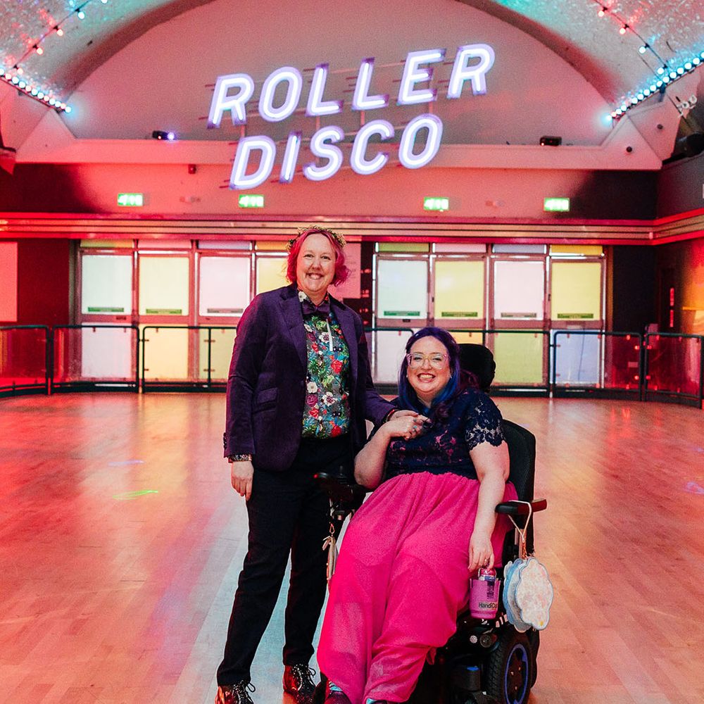 Bride in long pink wedding dress with purple bridal coverup on top with bride in purple velvet blazer and black suit trousers with purple bowtie, pink patterned pocket square and colourful patterned shirt in the roller disco of Margate Dreamland standing in front of large neon roller disco sign 