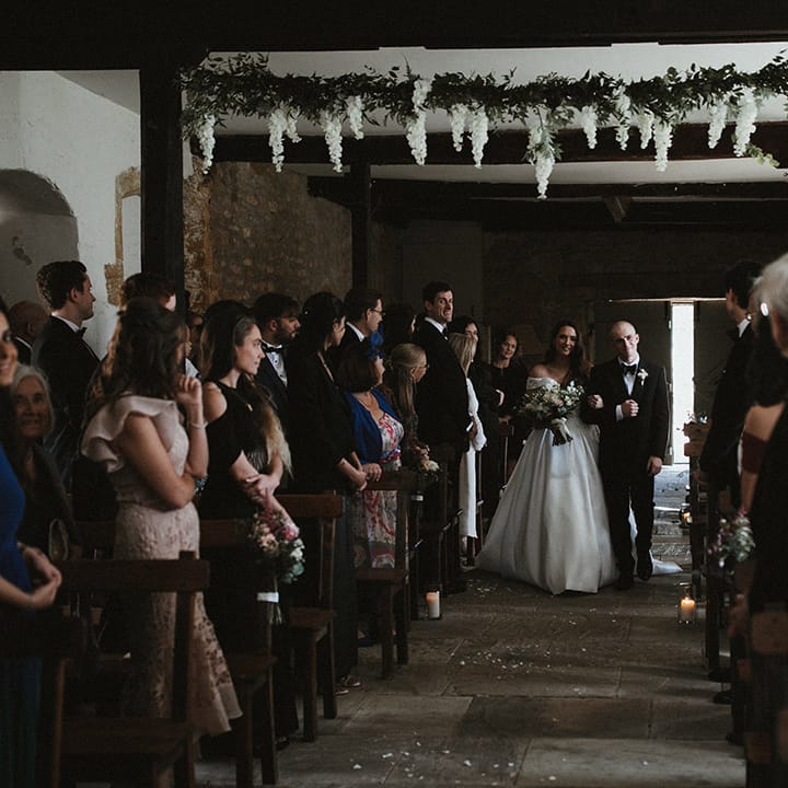 The bride in a Pronovias wedding dress is walked down he aisle by her brother in black tie 