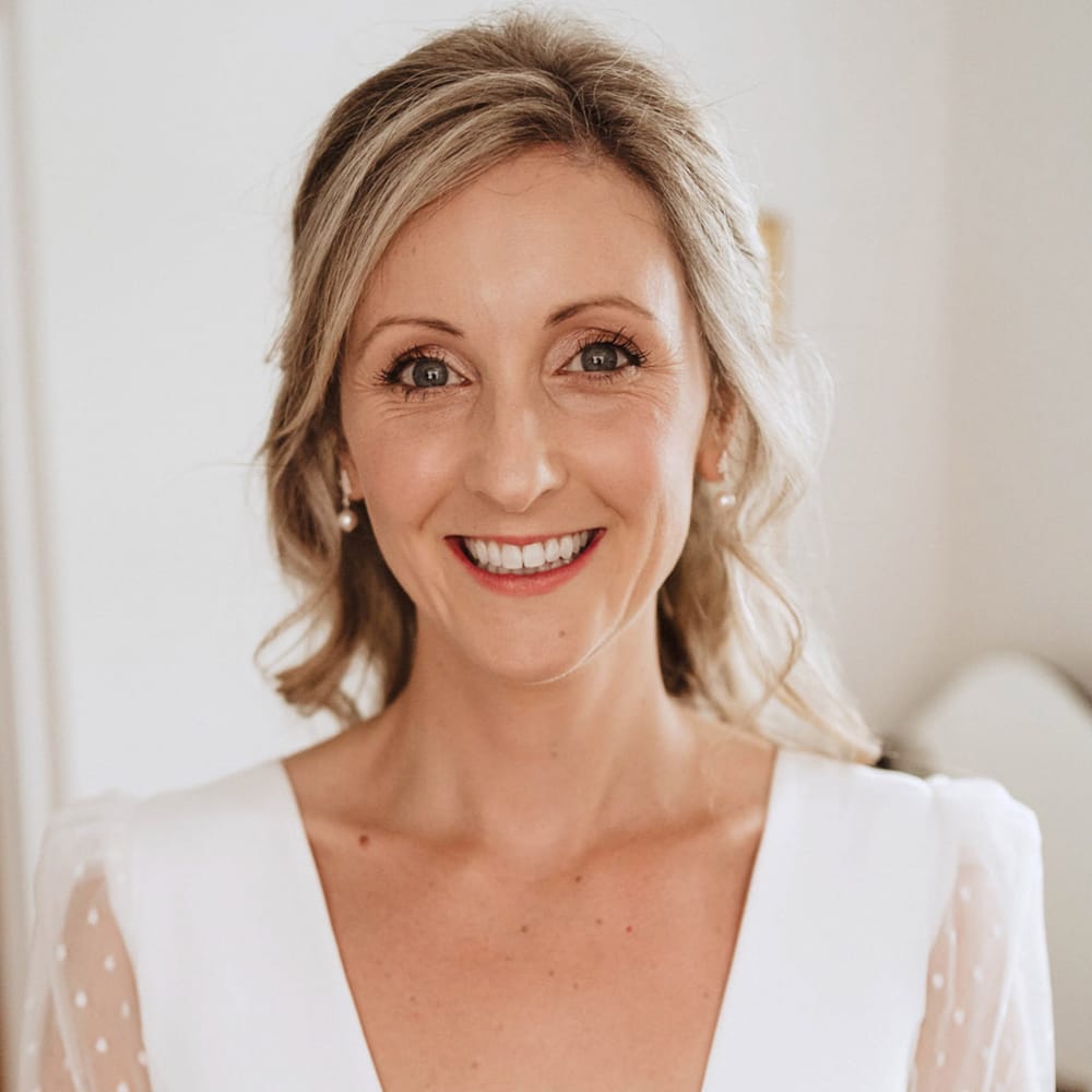 The bride smiles with her blonde hair in a half up half down style with pearl earrings and soft wedding makeup in a Sassi Holford wedding dress