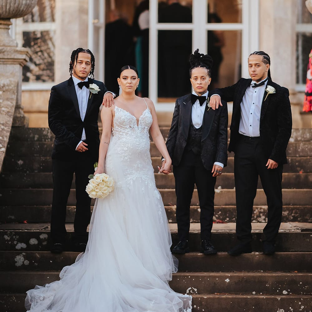 Bride in lace sleeveless v neck Enzoani wedding dress with tulle finish and church length veil holding hands with bride in black snakeskin print tuxedo with black bowtie and white rose boutonniere standing with bridal party in classic black tuxedos with white rose boutonnieres 