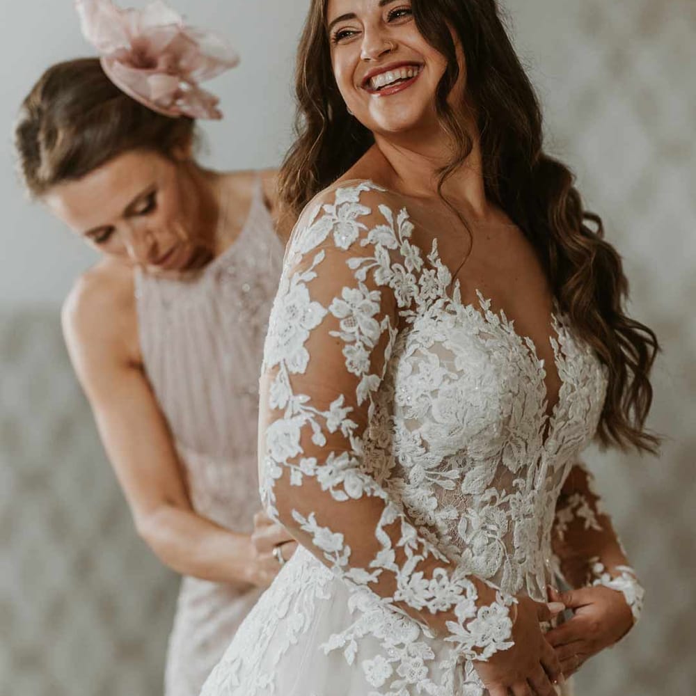Mother of the bride in dusky pink sleeveless dress and dusky pink fascinator helping bride with long sleeve lace illusion sleeve wedding dress with overlay 