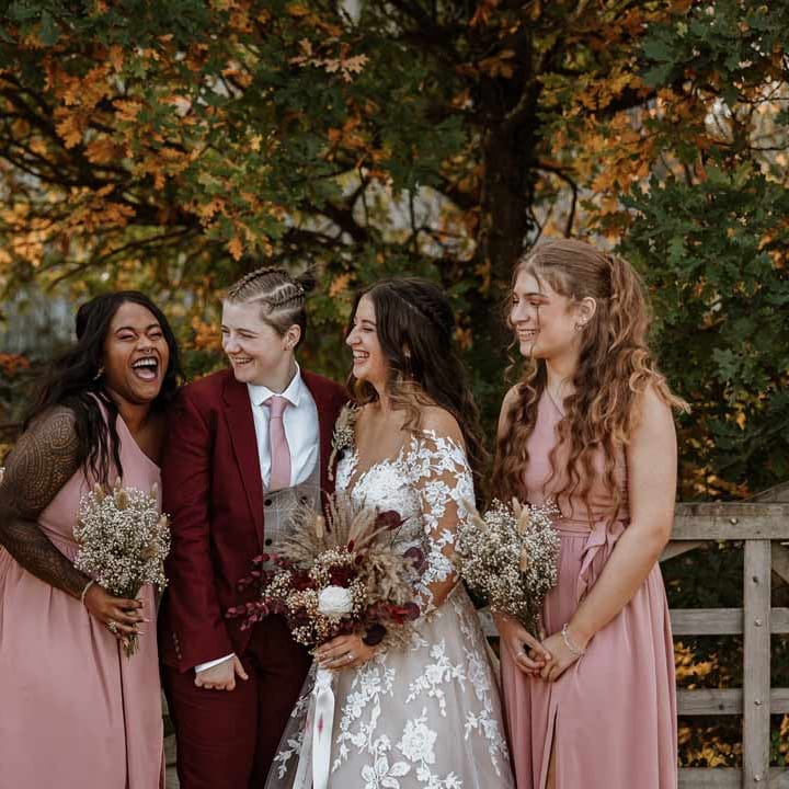 Bridesmaids in dusky pink one shoulder bridesmaid dresses standing with bride in maroon bridal suit with grey waistcoat, dusky pink tie and autumnal dried flower boutonniere and bride in long sleeve lace illusion sleeve wedding dress with overlay holding large autumnal boho dried flower bouquet 