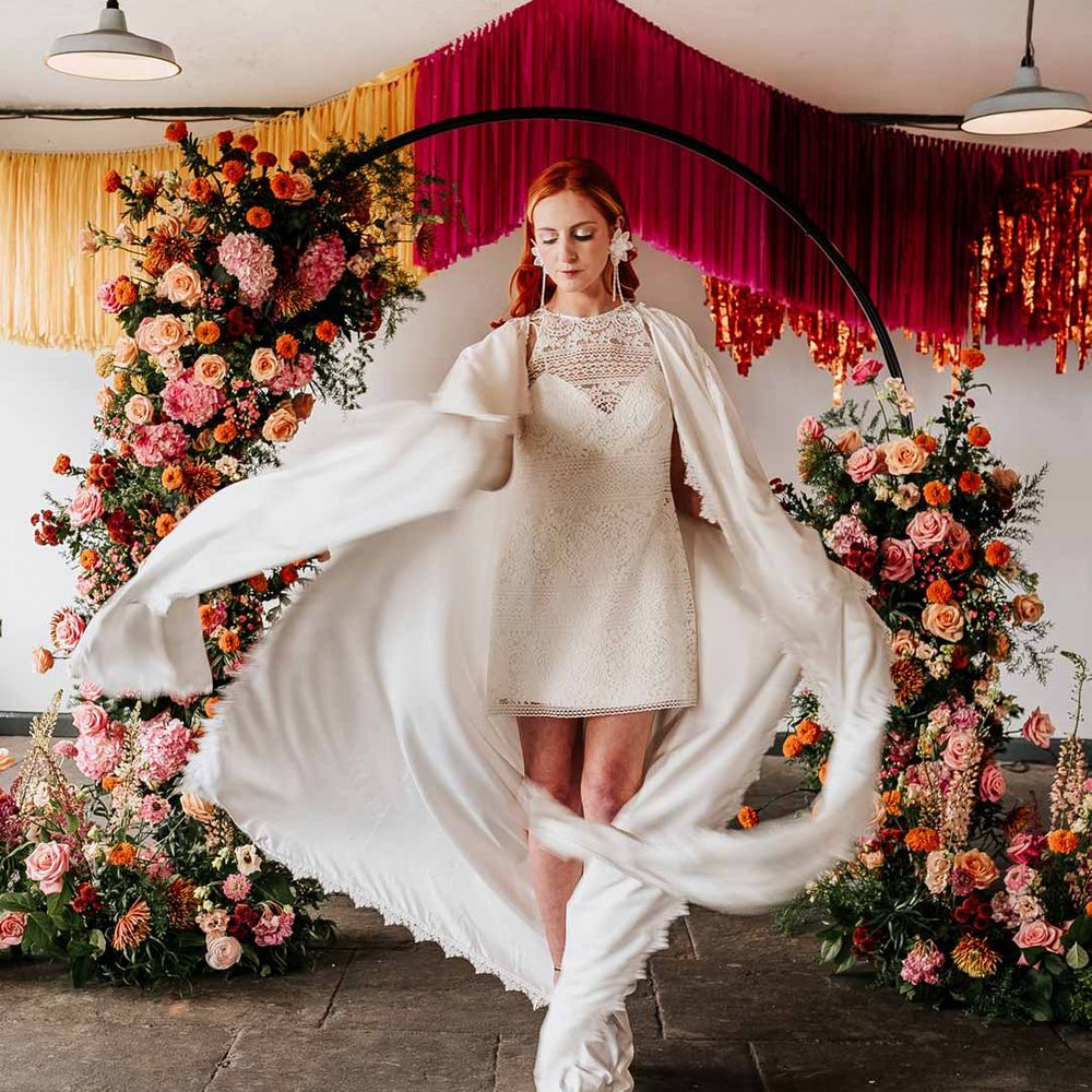 Bride in 70s inspired boatneck short lace wedding dress with detachable tulle puff sleeves, platform gold wedding heels and scallop hem bridal cape at Northern Monk Refectory Leeds standing by circular floral arch with vibrant flowers and foliage attached and pink, red, yellow and metallic wedding streamers behind