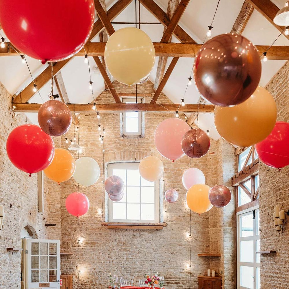 Merriscourt wedding venue reception room with white wooden chairs, metallic pink, white, orange and yellow balloons, wedding fairy lights decorations and wooden table with orange table runner and mixed seasonal flower arrangements 