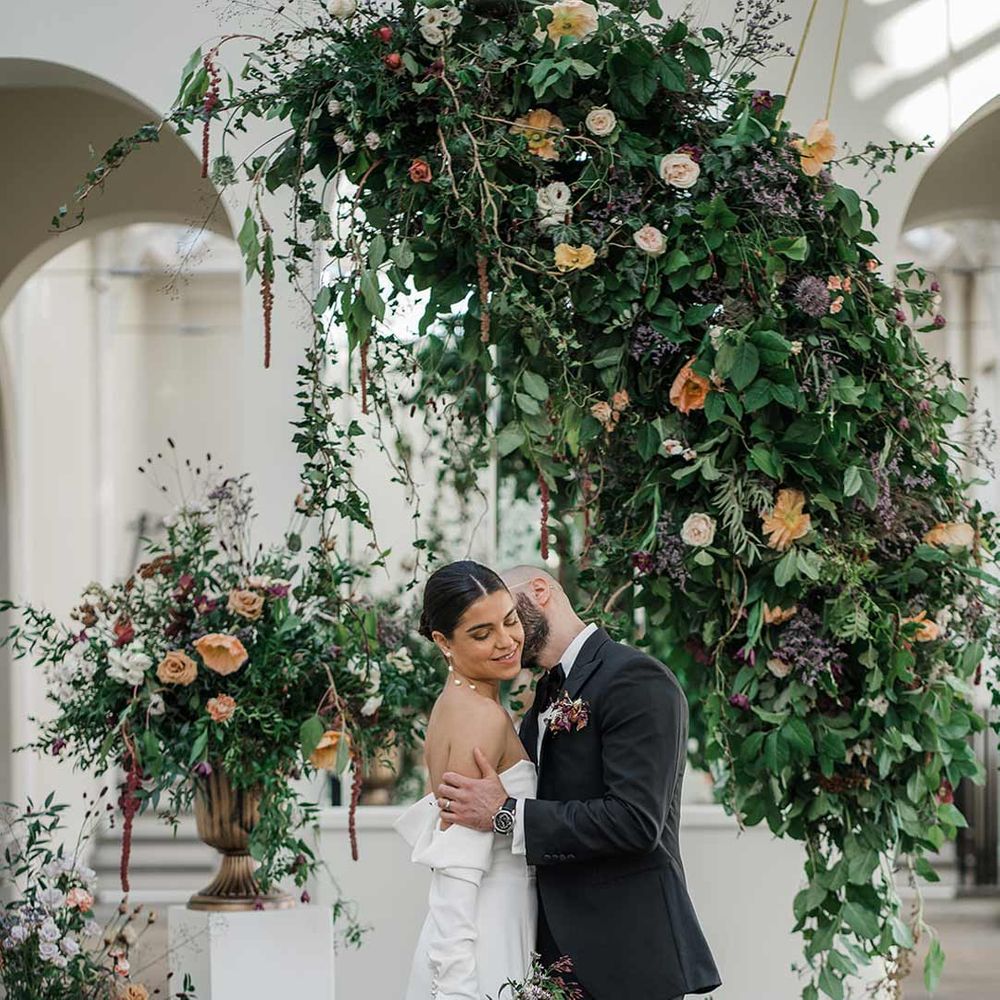 Groom in classic black tuxedo with bowtie and wildflower boutonniere embracing bride in long sleeve off the shoulder wedding dress with pearl button detailing and a side slit holding dried wildflower bouquet surrounded by suspended wildflower and foliage arrangements in large gold vases at Buxted Park